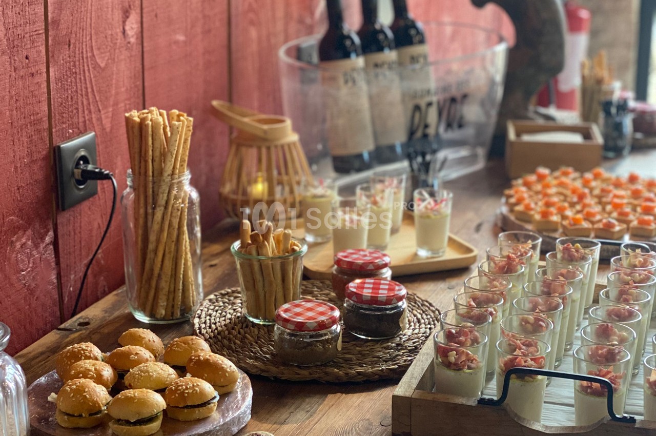 Buffet avec mini-burgers, verrines, gressins, pots de confiture et bouteilles de vin sur une table en bois.