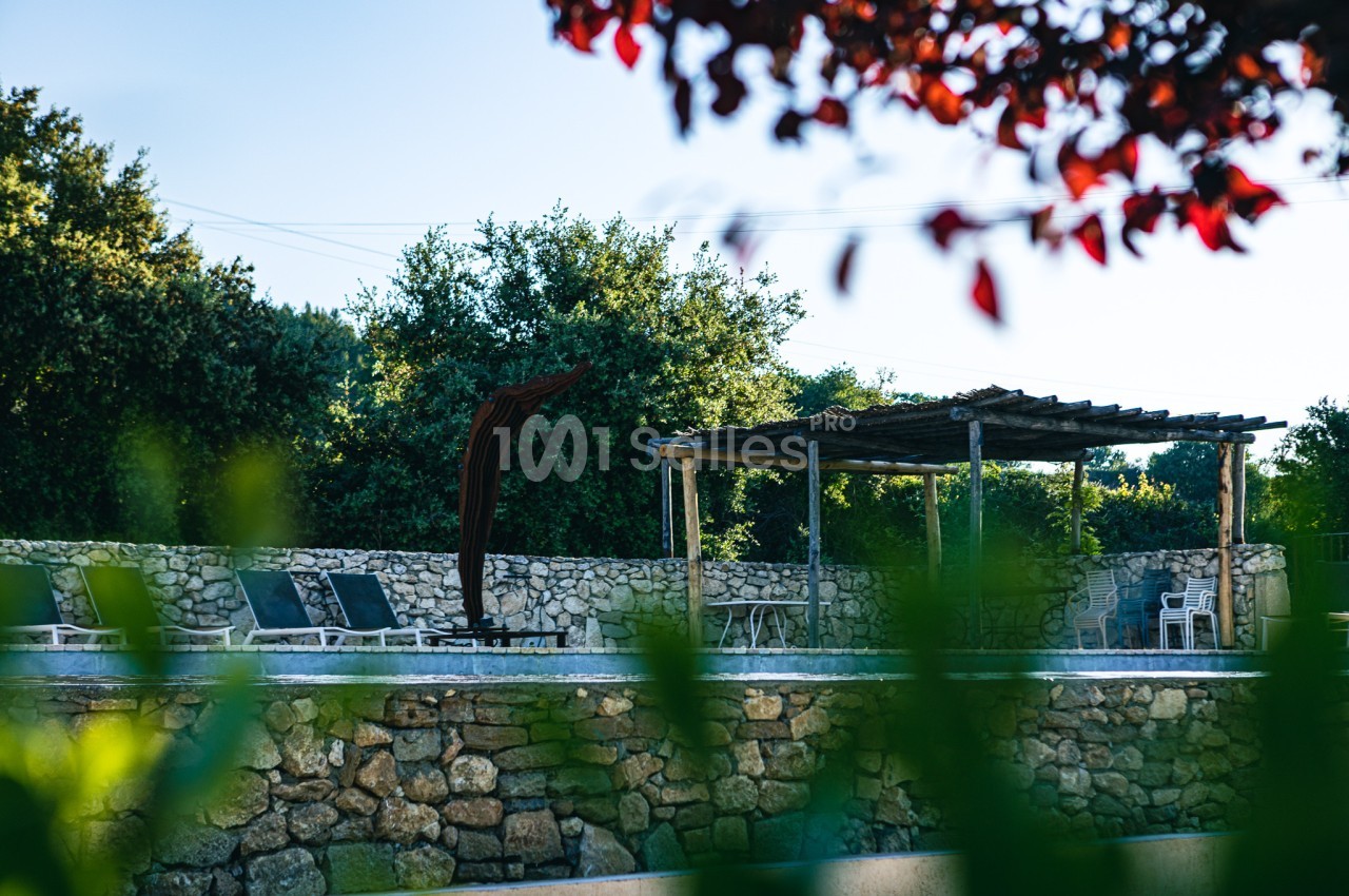 Terrasse avec pergola en bois, chaises longues et piscine entourée de murs en pierre, vue à travers des feuillages.
