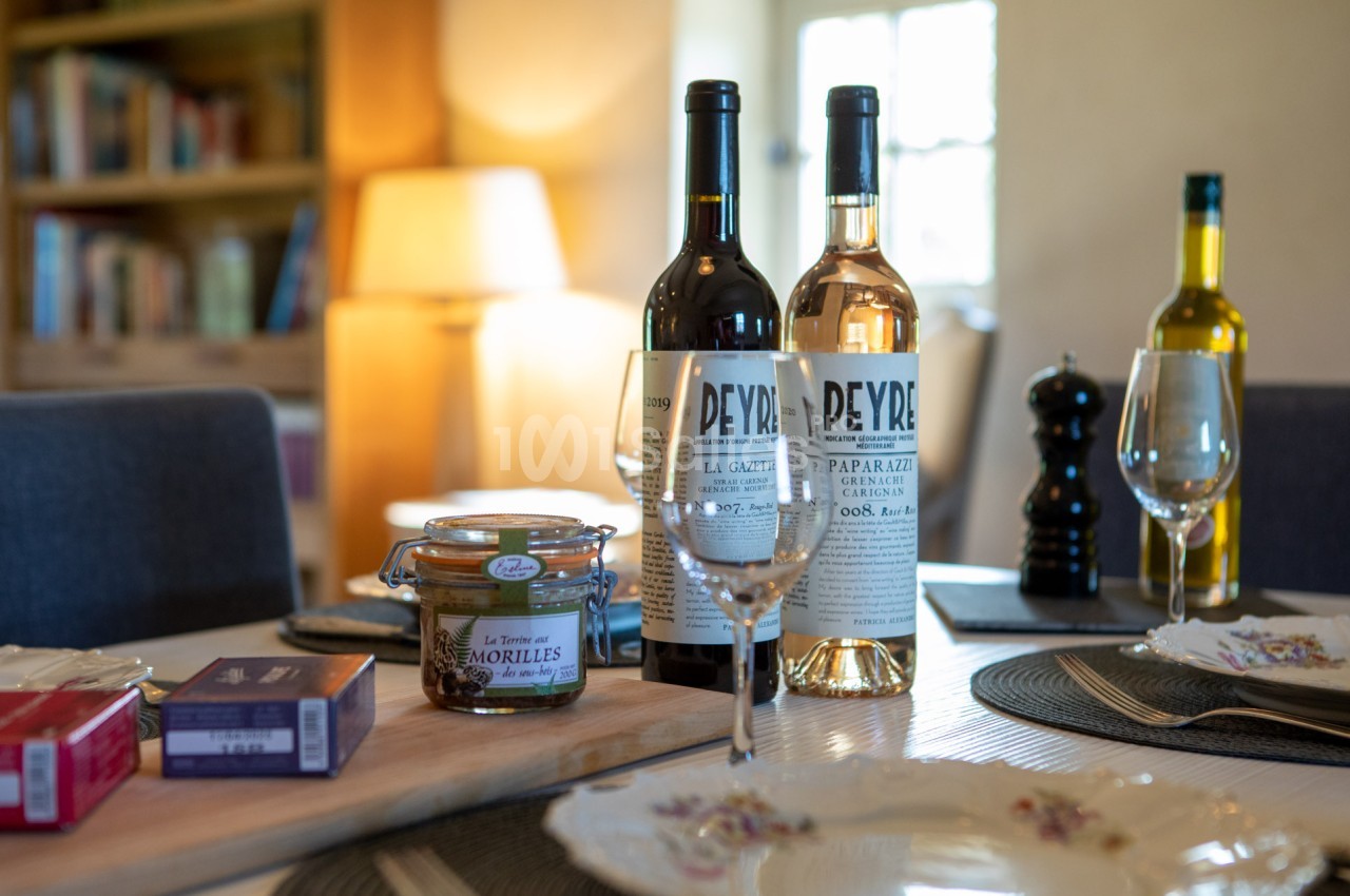 Bouteilles de vin, verres, pot de confiture et assiettes disposés sur une table dressée dans un intérieur chaleureux.