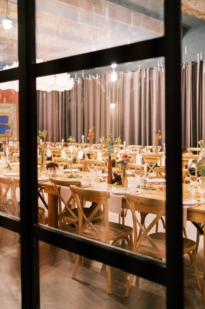 Salle de réception décorée avec des tables en bois, chaises assorties et centres de table fleuris, vue à travers une fenêtre.