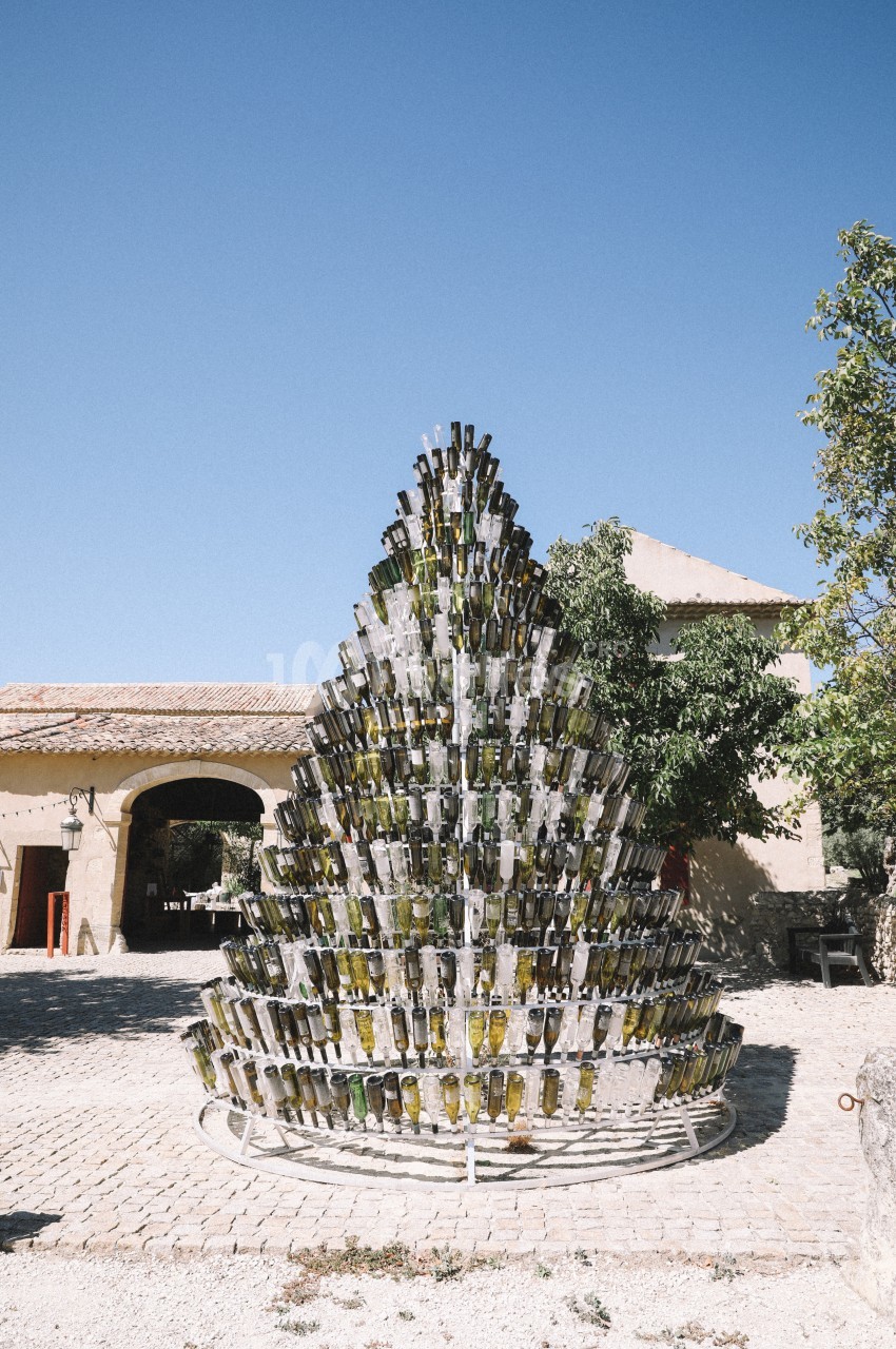 Structure en forme de sapin composée de bouteilles en verre, installée sur une place pavée sous un ciel bleu.