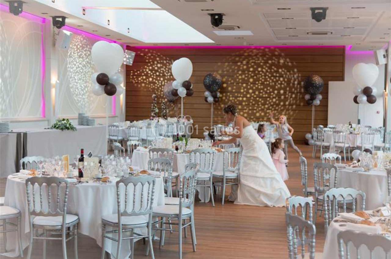 Salle de réception décorée pour un mariage, avec tables dressées, ballons et une mariée ajustant les détails.