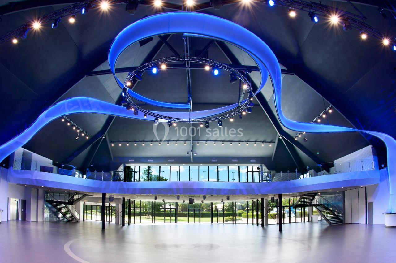 Intérieur spacieux d'une salle moderne avec éclairage bleu, mezzanine vitrée et grandes baies vitrées donnant sur un jardin.