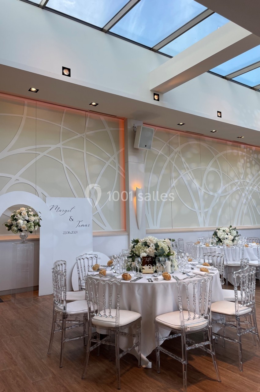 Salle de réception lumineuse avec tables rondes dressées, décor floral blanc et mur orné de motifs abstraits.