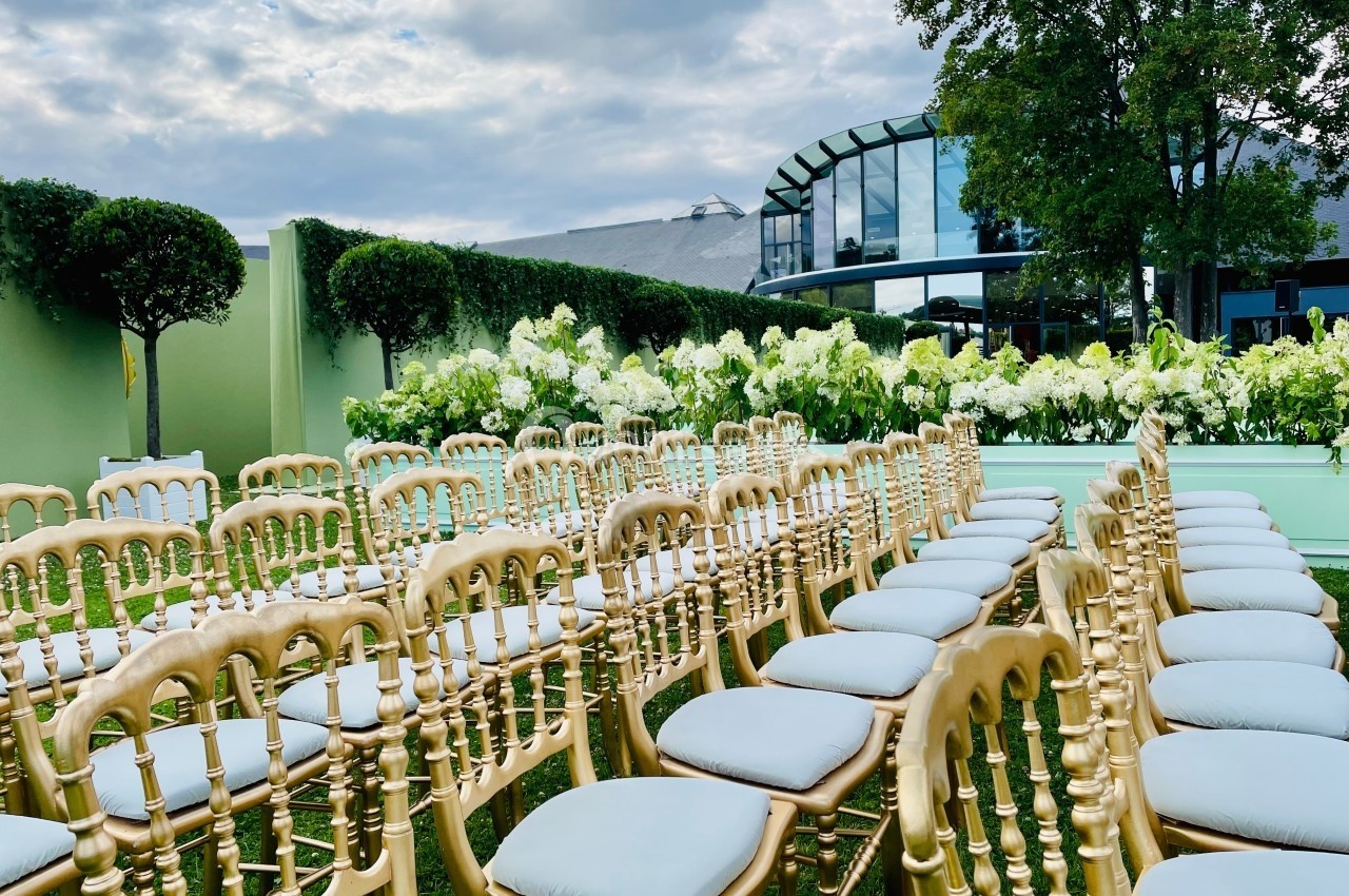 Chaises dorées alignées en extérieur, entourées de fleurs blanches et de haies, devant un bâtiment moderne vitré.