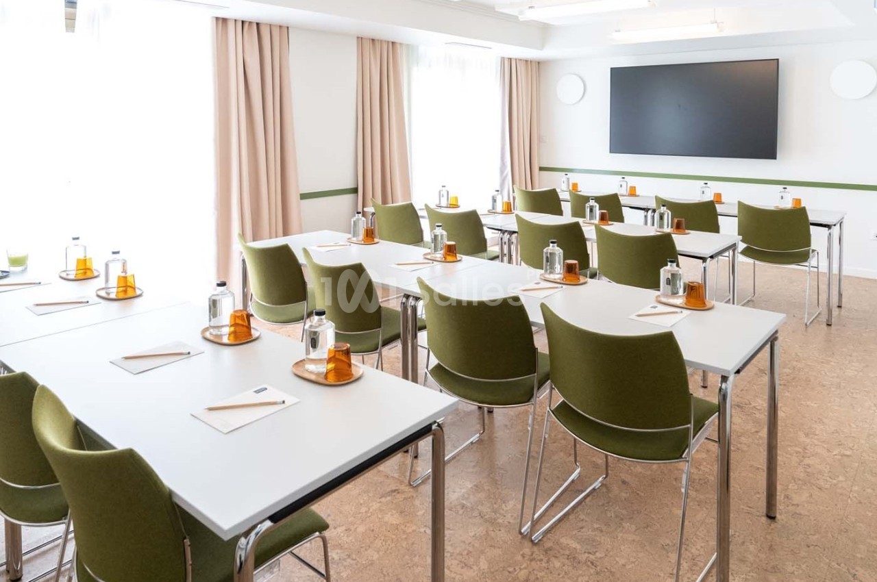Salle de réunion lumineuse avec des tables blanches, des chaises vertes et un écran noir fixé au mur.
