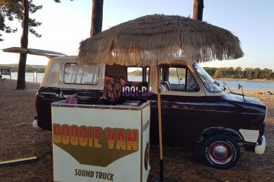 Un DJ mixe à l'extérieur depuis un stand aménagé dans un véhicule, avec des tables et des personnes en arrière-plan.