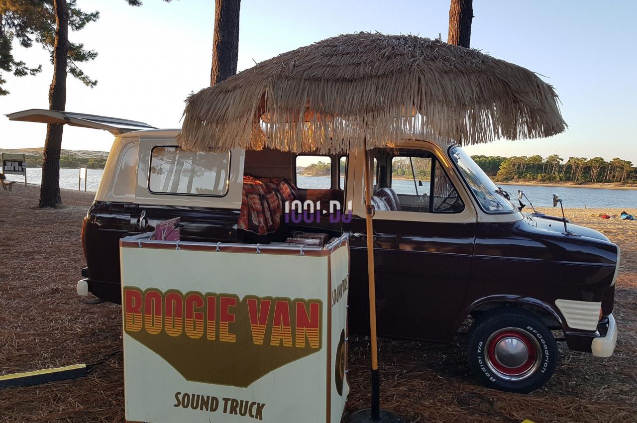 Un van vintage aménagé avec un comptoir et un parasol en paille, stationné près d'un lac entouré de pins.