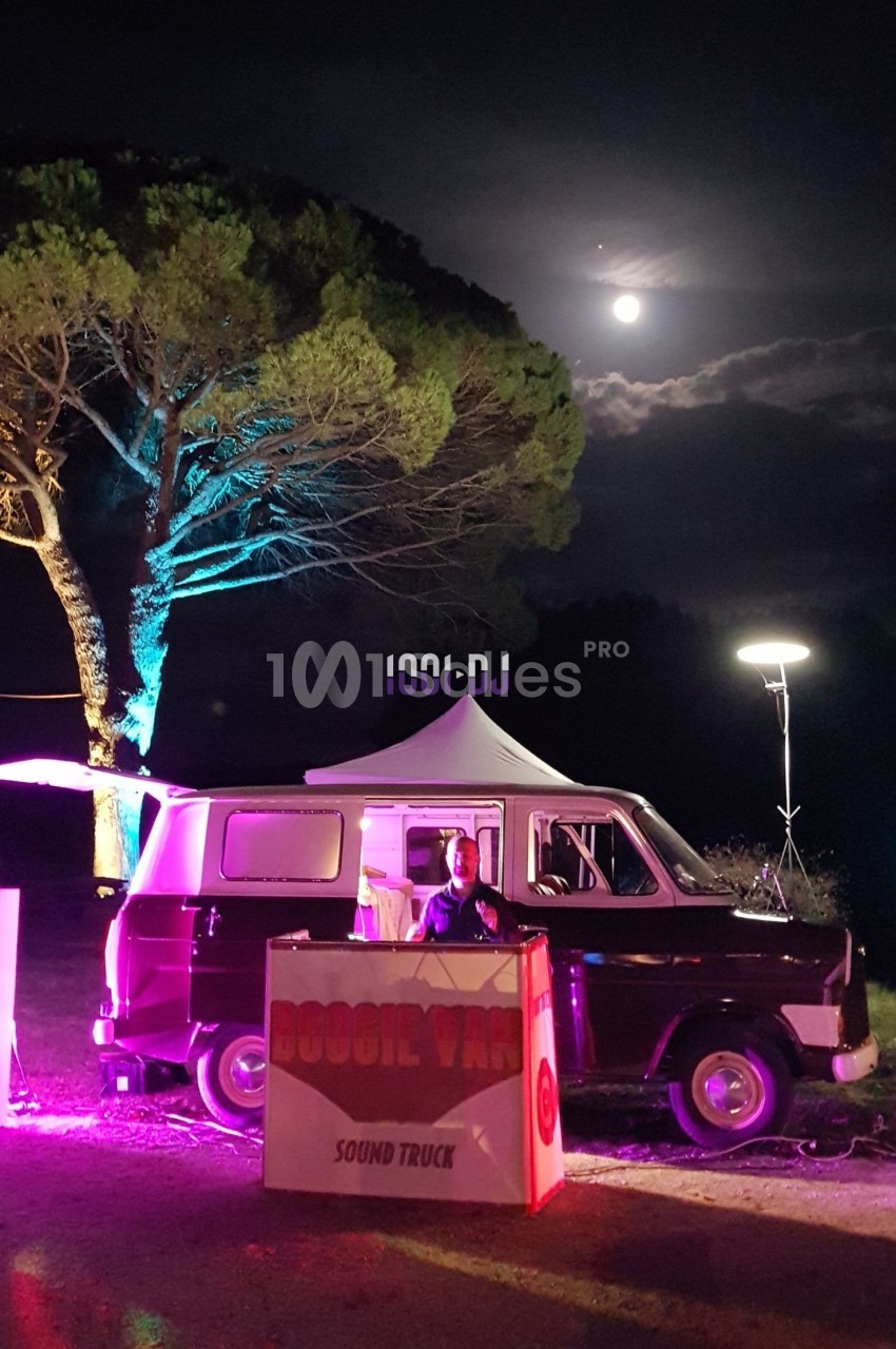Un DJ mixe devant un van éclairé, entouré de haut-parleurs, sous un ciel nocturne avec une pleine lune.