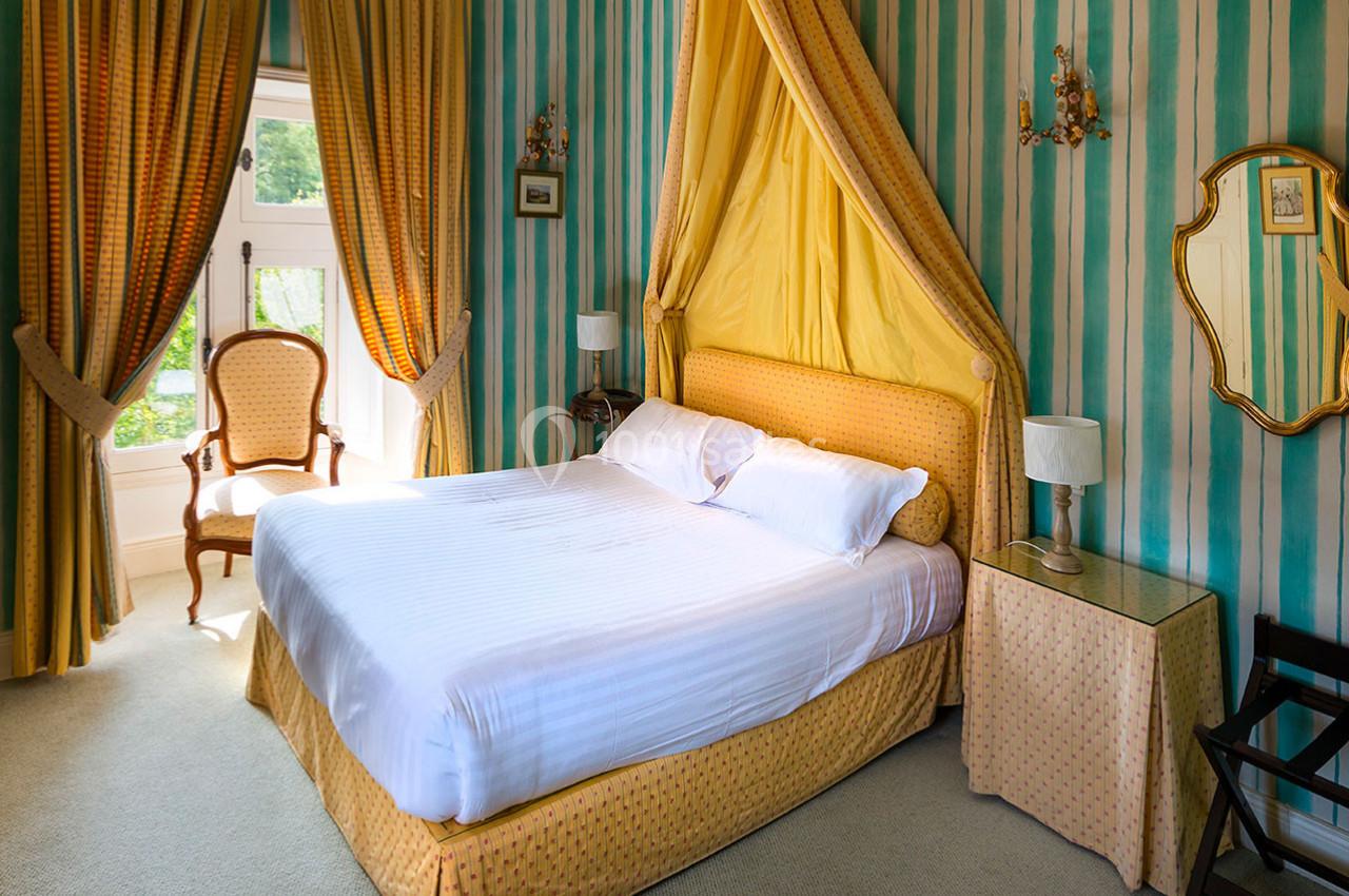 Lit double avec baldaquin jaune dans une chambre aux murs rayés vert et jaune, éclairée par une fenêtre.