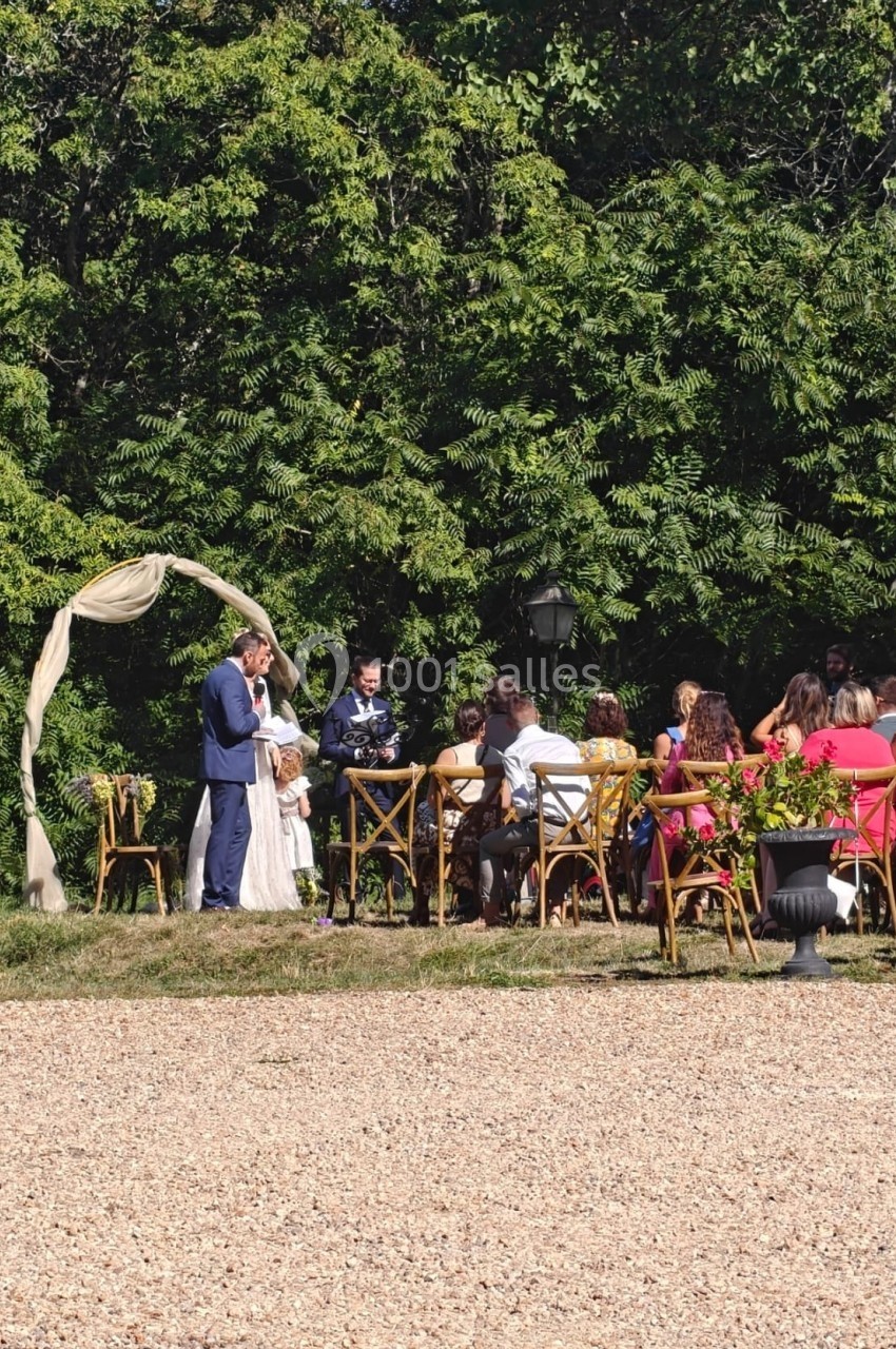 Un couple échange ses vœux sous une arche décorée, entouré d'invités assis en extérieur près d'arbres.