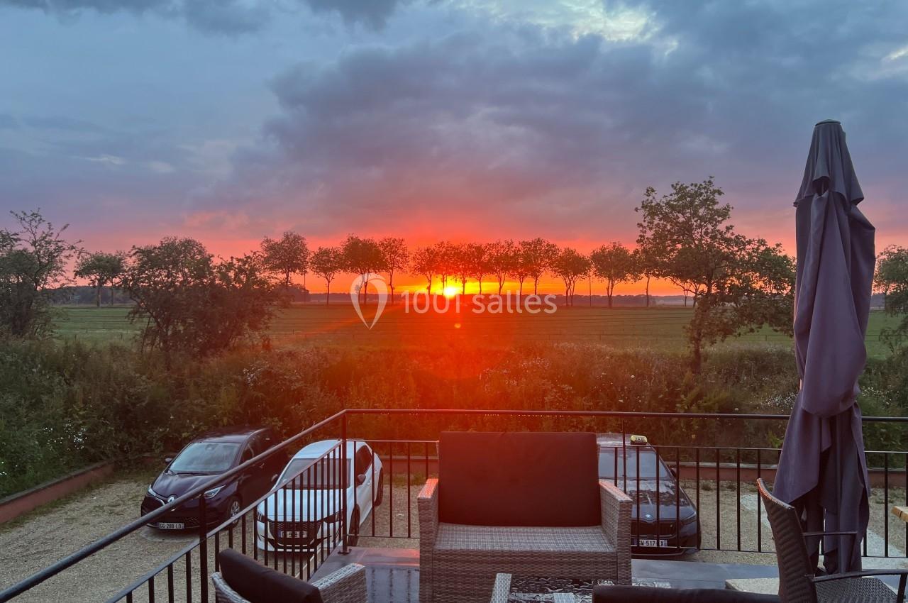 Coucher de soleil rougeoyant derrière une rangée d'arbres, vu depuis une terrasse avec mobilier et voitures garées.