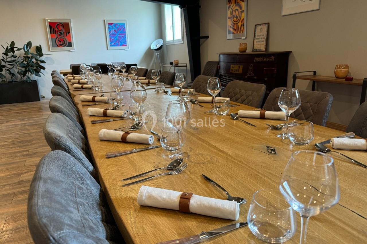Grande table en bois dressée avec couverts, verres et serviettes, entourée de chaises dans une salle décorée de tableaux.