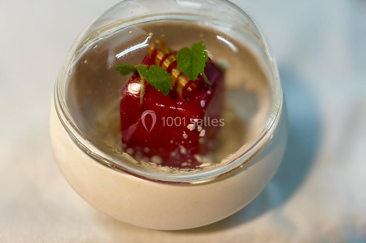 Cube de dessert rouge décoré de feuilles vertes et d'éléments dorés, présenté dans un bol transparent sur une nappe blanche.