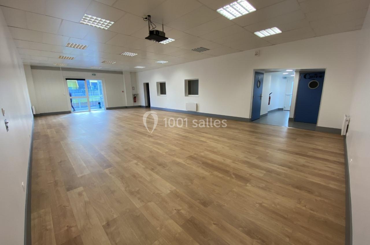 Salle vide avec parquet clair, murs blancs, plafond à dalles et éclairage fluorescent, portes et fenêtres visibles.
