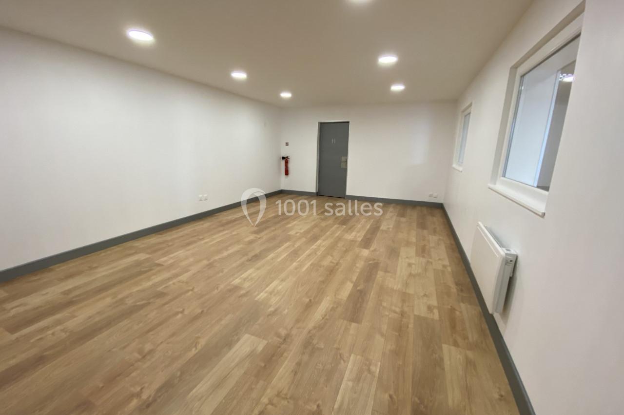 Salle vide avec parquet en bois clair, murs blancs, éclairage encastré au plafond et une porte grise au fond.