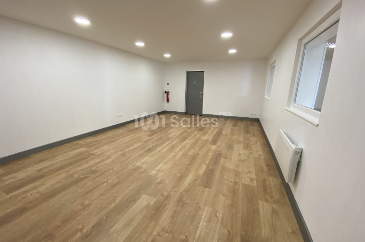 Salle vide avec parquet clair, murs blancs, éclairage encastré au plafond et une porte grise au fond.