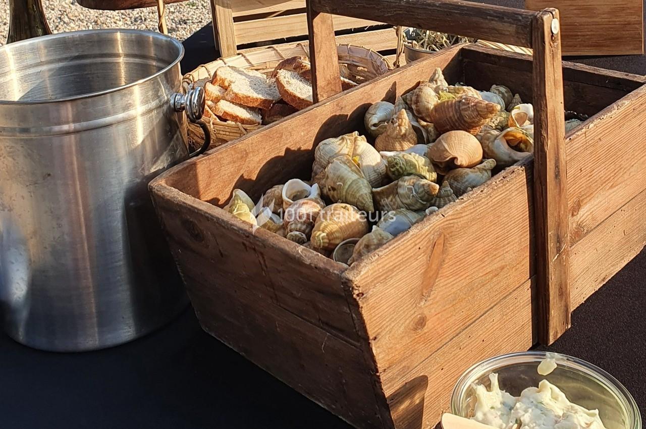 Cagette en bois remplie de bulots cuits, accompagnée de pain, d'une sauce et d'un seau métallique en arrière-plan.