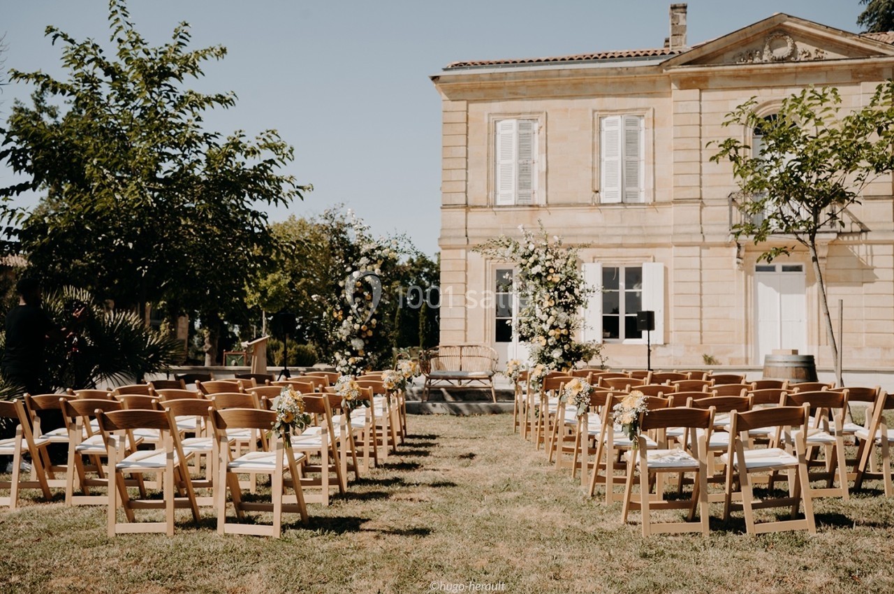 Chaises en bois disposées en rangées devant une maison en pierre, décorées pour une cérémonie en extérieur.