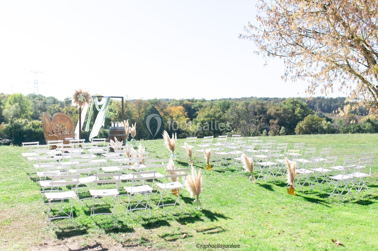 Chaises blanches disposées en rangées sur une pelouse pour une cérémonie en plein air, avec arche décorée et paysage boisé.