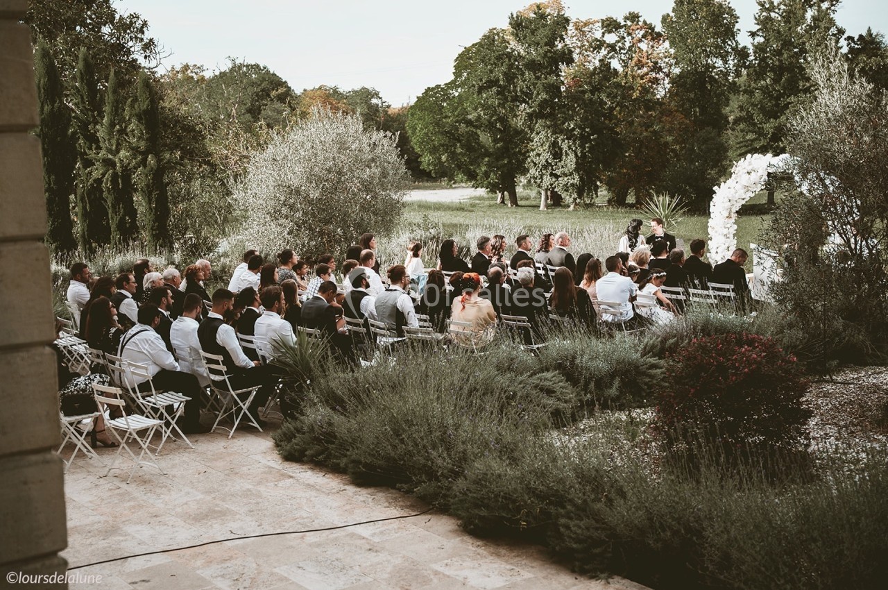 Cérémonie en plein air avec des invités assis, entourés de verdure et d'une arche florale blanche.