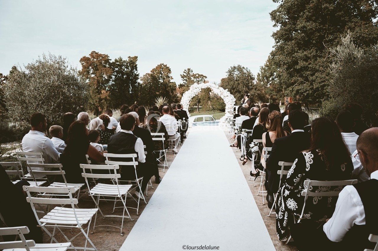 Allée blanche menant à une arche fleurie lors d'une cérémonie de mariage en plein air, entourée d'invités assis.