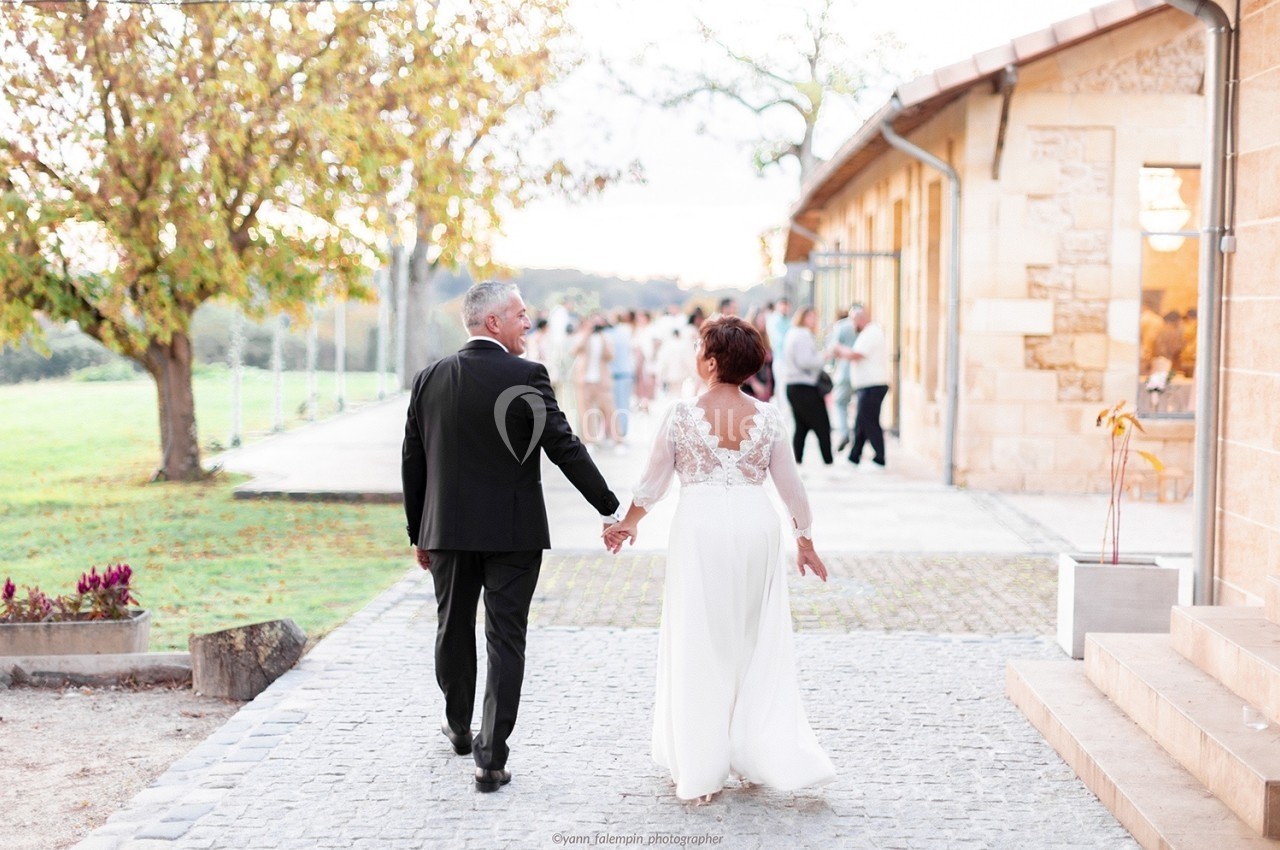 Un couple en tenue de mariage marche main dans la main sur une allée pavée près d'un bâtiment en pierre.