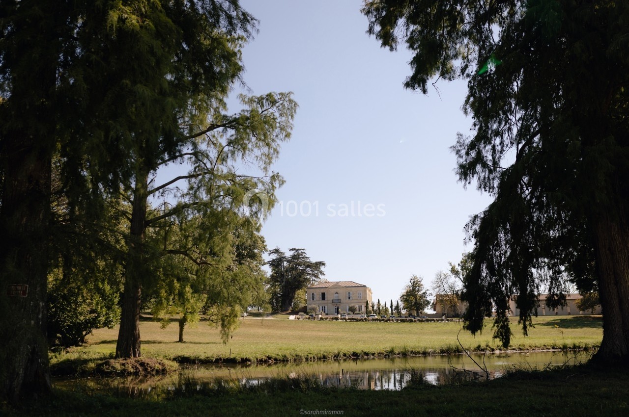 Vue d'une grande maison entourée d'arbres et d'une pelouse, avec un petit plan d'eau au premier plan.