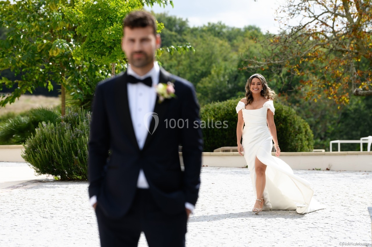 Une mariée souriante s'approche d'un homme en costume, flou au premier plan, dans un cadre extérieur verdoyant.