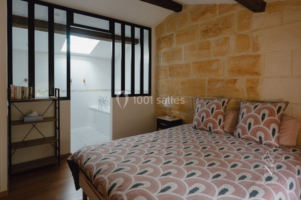 Chambre avec lit double, mur en pierre, étagère, et vue partielle sur une salle de bain séparée par une cloison vitrée.