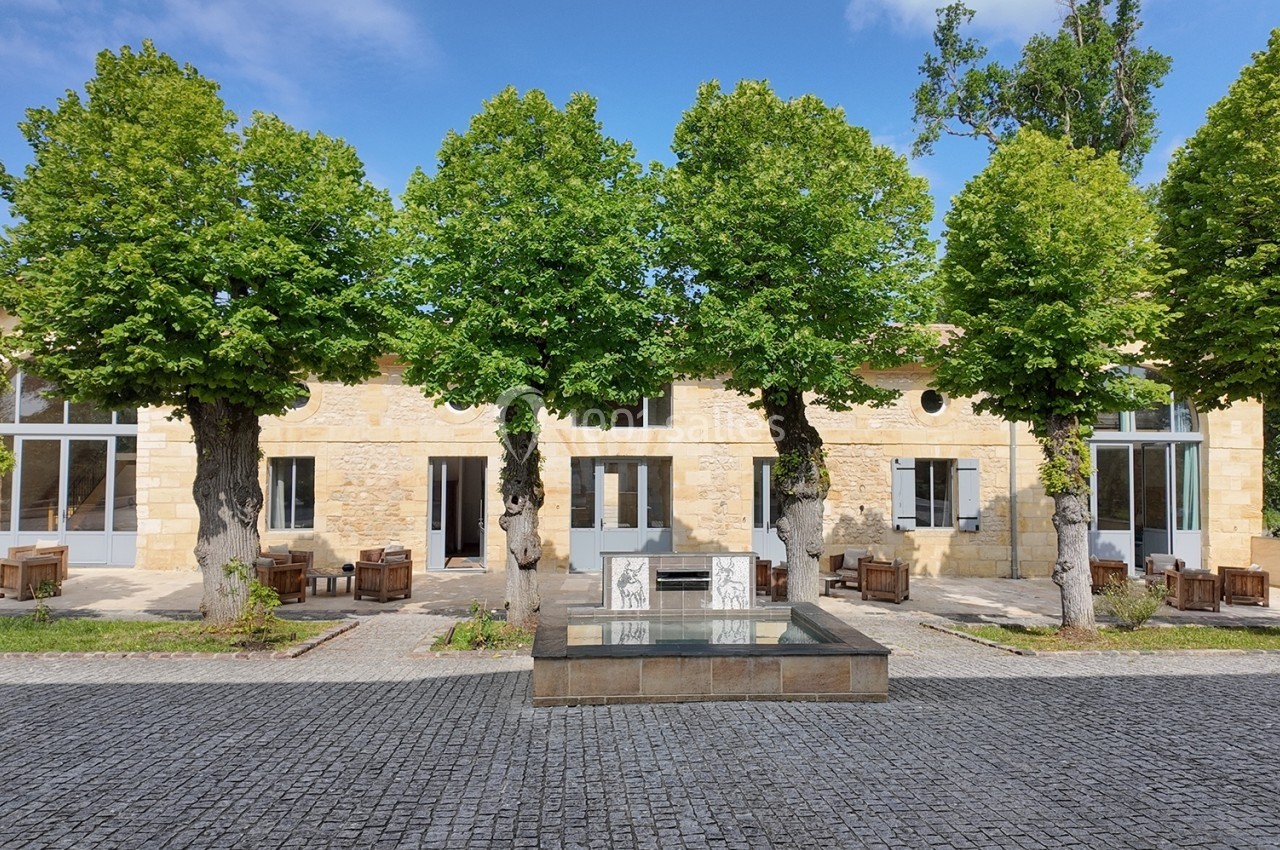 Façade en pierre d'un bâtiment avec des arbres alignés devant et une fontaine centrale sur une place pavée.