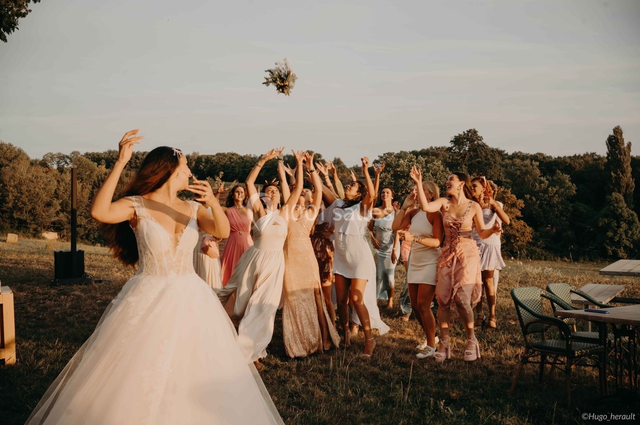 Une mariée lance son bouquet à un groupe de femmes souriantes dans un champ ensoleillé.