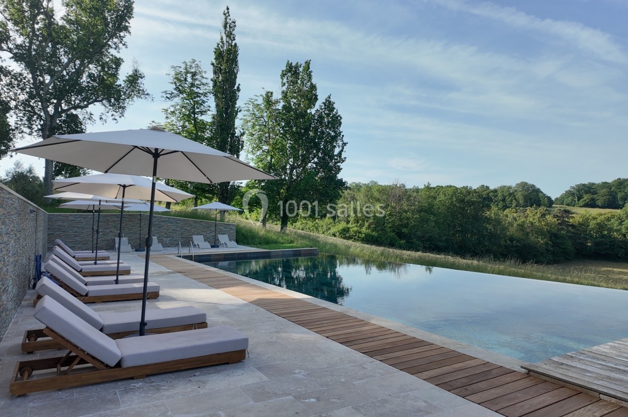 Chaises longues et parasols alignés au bord d'une piscine à débordement, entourée de verdure et d'un mur en pierre.