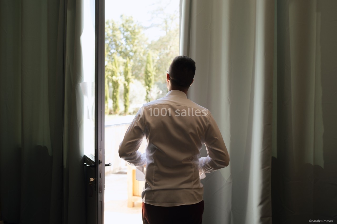 Un homme en chemise blanche regarde à l'extérieur depuis une porte ouverte, entourée de rideaux.