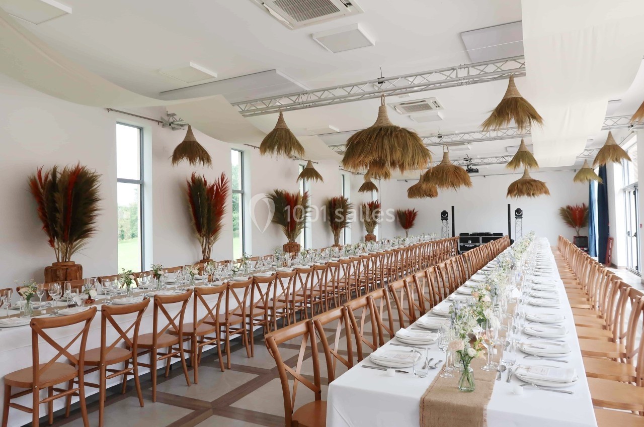 Salle de réception décorée avec des tables longues, nappes blanches, chaises en bois et suspensions en paille.
