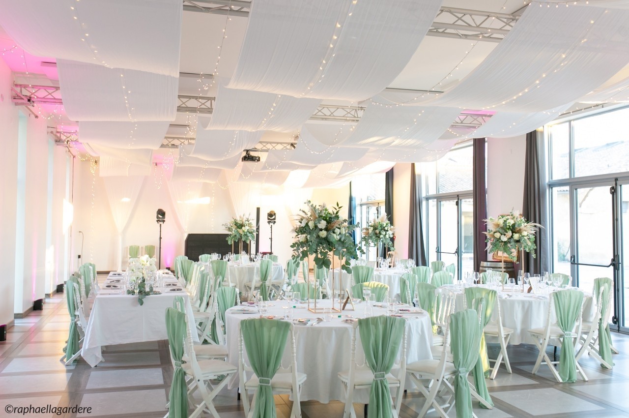 Salle de réception décorée avec des tables rondes, nappes blanches, chaises ornées de tissu vert et éclairage tamisé.