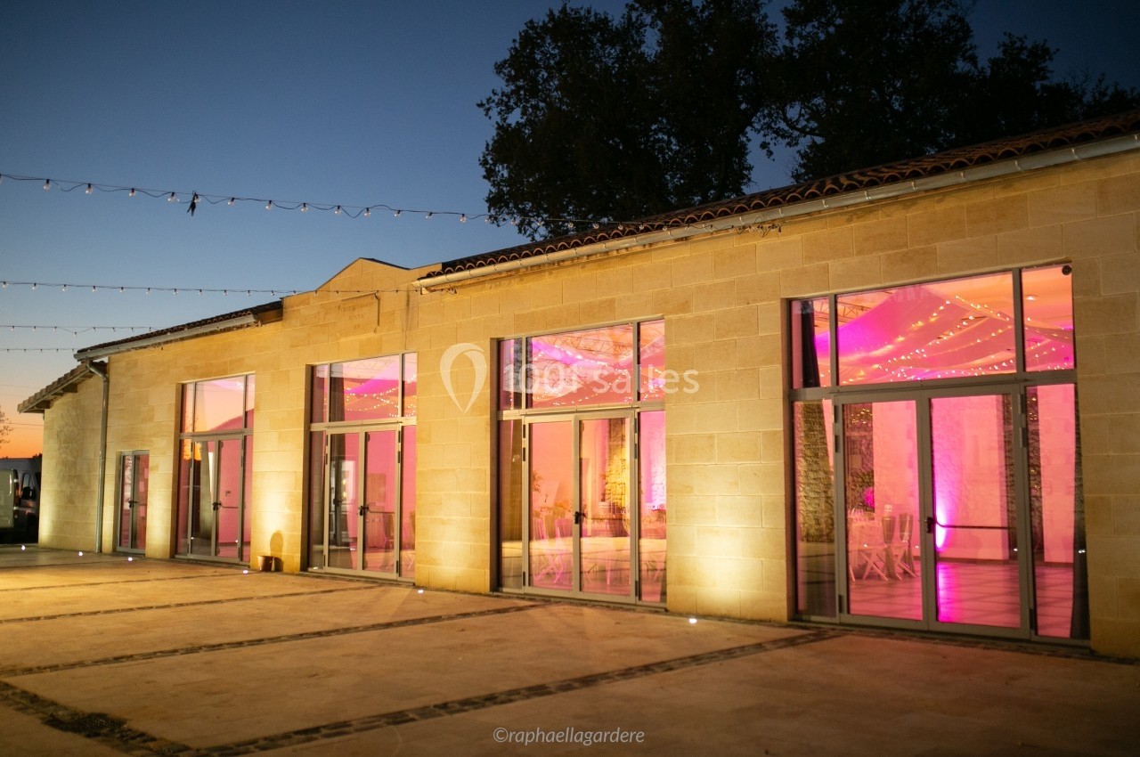 Façade d'un bâtiment en pierre avec de grandes baies vitrées illuminées par des lumières roses à la tombée de la nuit.