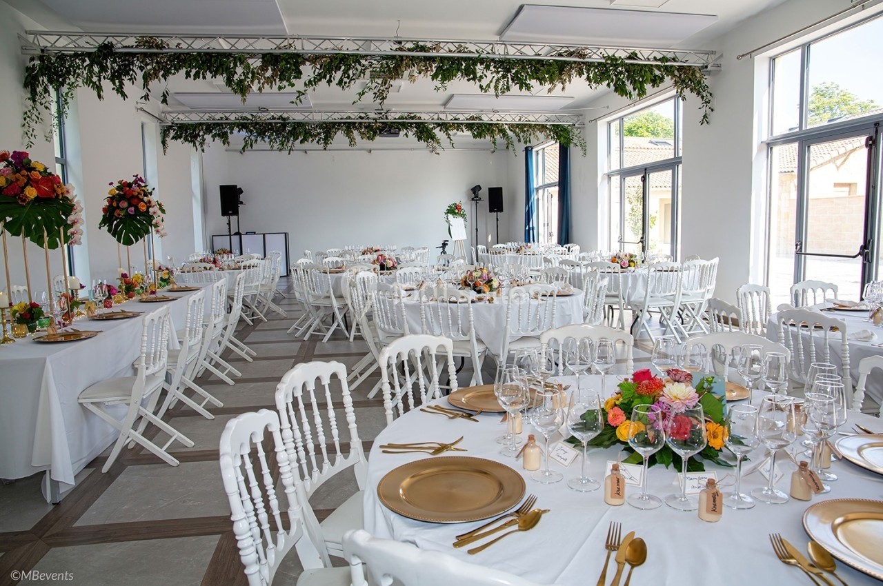 Salle de réception lumineuse décorée avec des guirlandes végétales, des tables rondes et des chaises blanches élégantes.