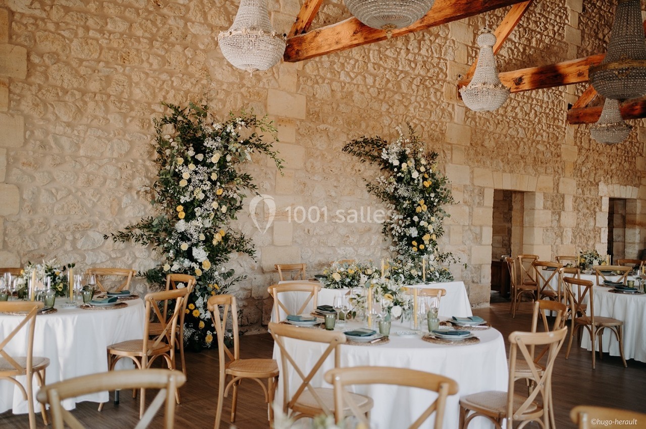 Salle de réception avec murs en pierre, tables rondes décorées et arrangements floraux blancs et jaunes.