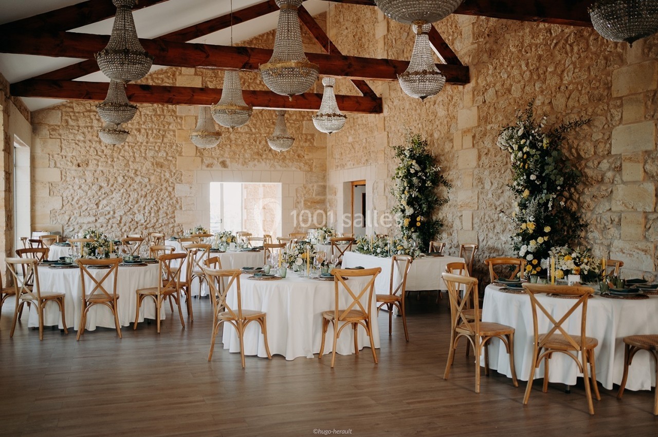 Salle de réception lumineuse avec murs en pierre, tables rondes décorées et chandeliers suspendus.