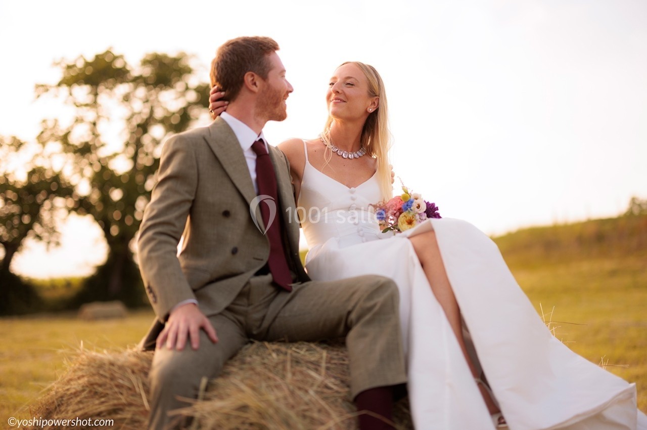 Un couple assis sur une botte de foin dans un champ, la femme en robe blanche tenant un bouquet de fleurs.