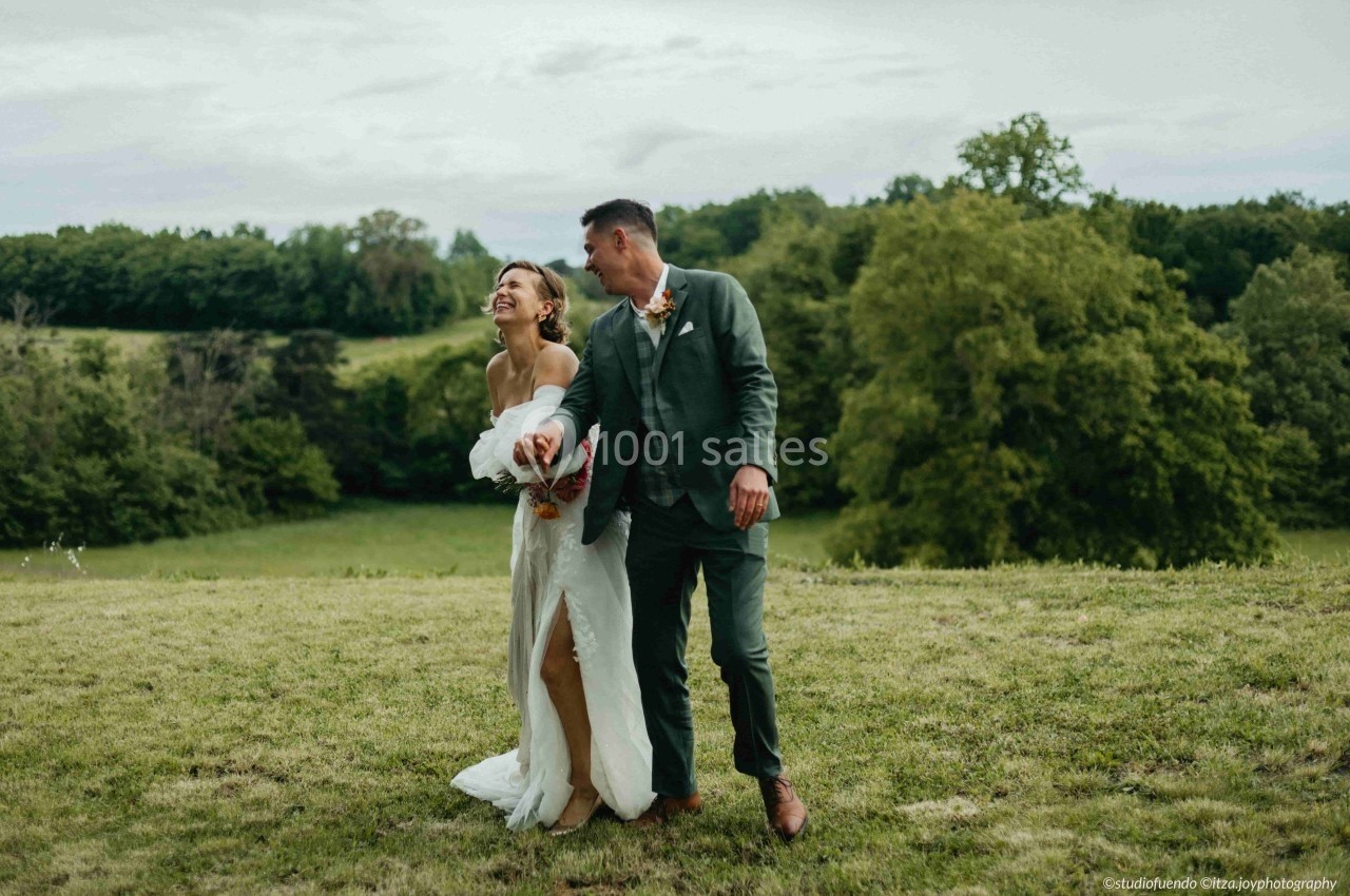 Un couple souriant marche dans un champ verdoyant sous un ciel nuageux.