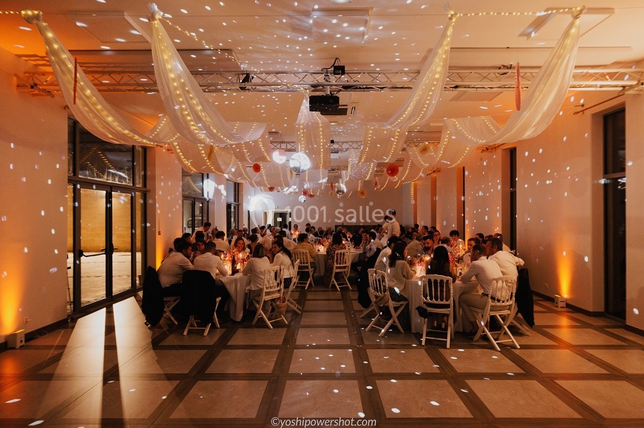 Salle de réception décorée de voilages lumineux, avec des invités assis à des tables rondes lors d'un événement.