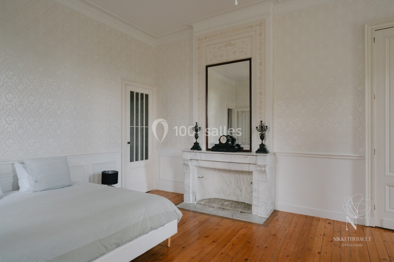 Chambre lumineuse avec lit, cheminée en marbre blanc, grand miroir et parquet en bois clair.
