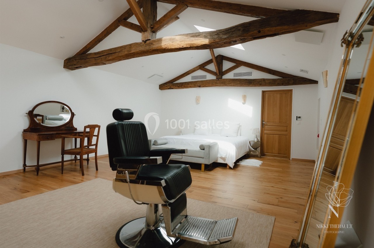 Chambre lumineuse avec poutres apparentes, lit double, coiffeuse en bois et fauteuil de barbier sur parquet.