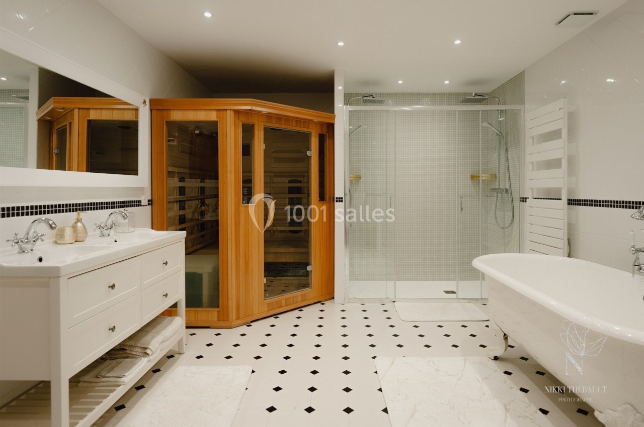 Salle de bain moderne avec sauna en bois, baignoire sur pieds, double vasque et douche vitrée sur carrelage noir et blanc.