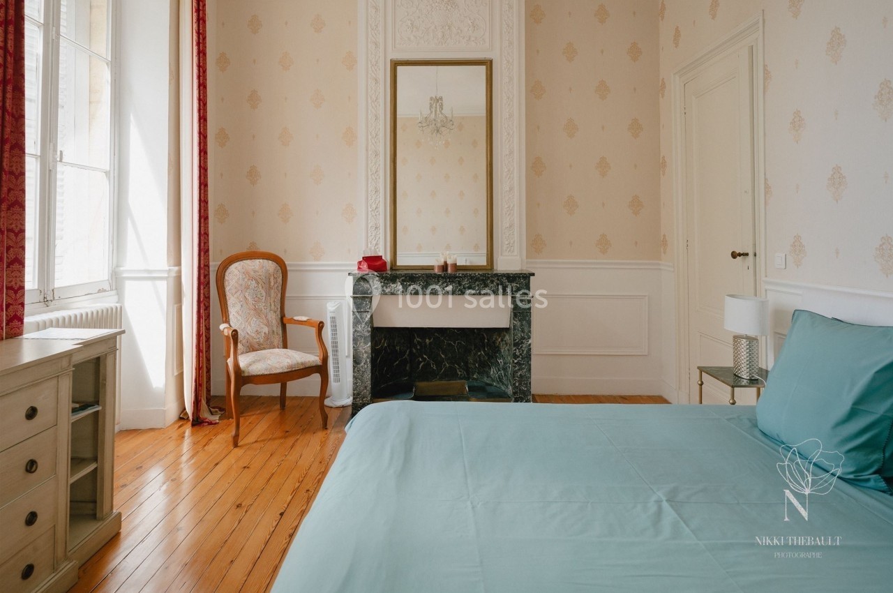 Chambre lumineuse avec lit double, cheminée en marbre, fauteuil en bois et grandes fenêtres avec rideaux rouges.