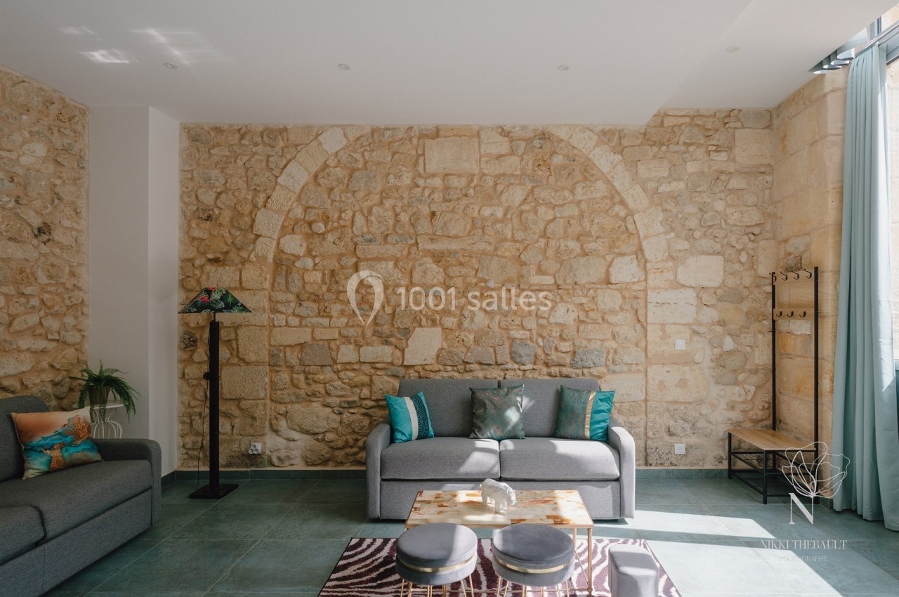 Salon lumineux avec murs en pierre apparente, canapé gris, table basse et grandes fenêtres laissant entrer la lumière.