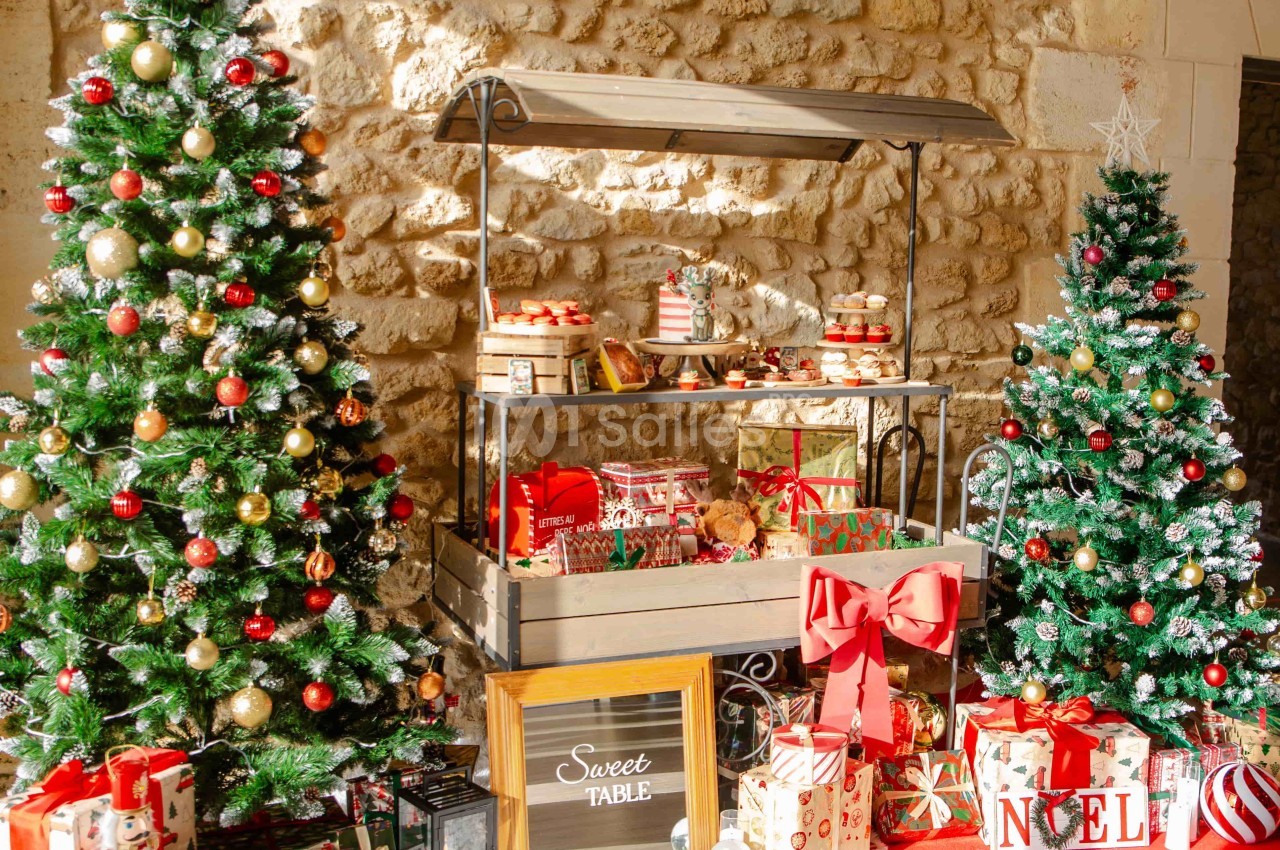 Deux sapins décorés entourent une table garnie de cadeaux et de douceurs dans un décor en pierre.