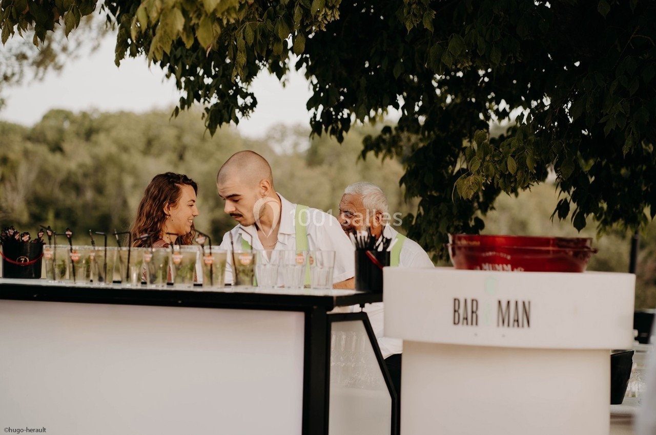 Trois personnes derrière un bar extérieur, entourées de verres et d'arbres en arrière-plan.